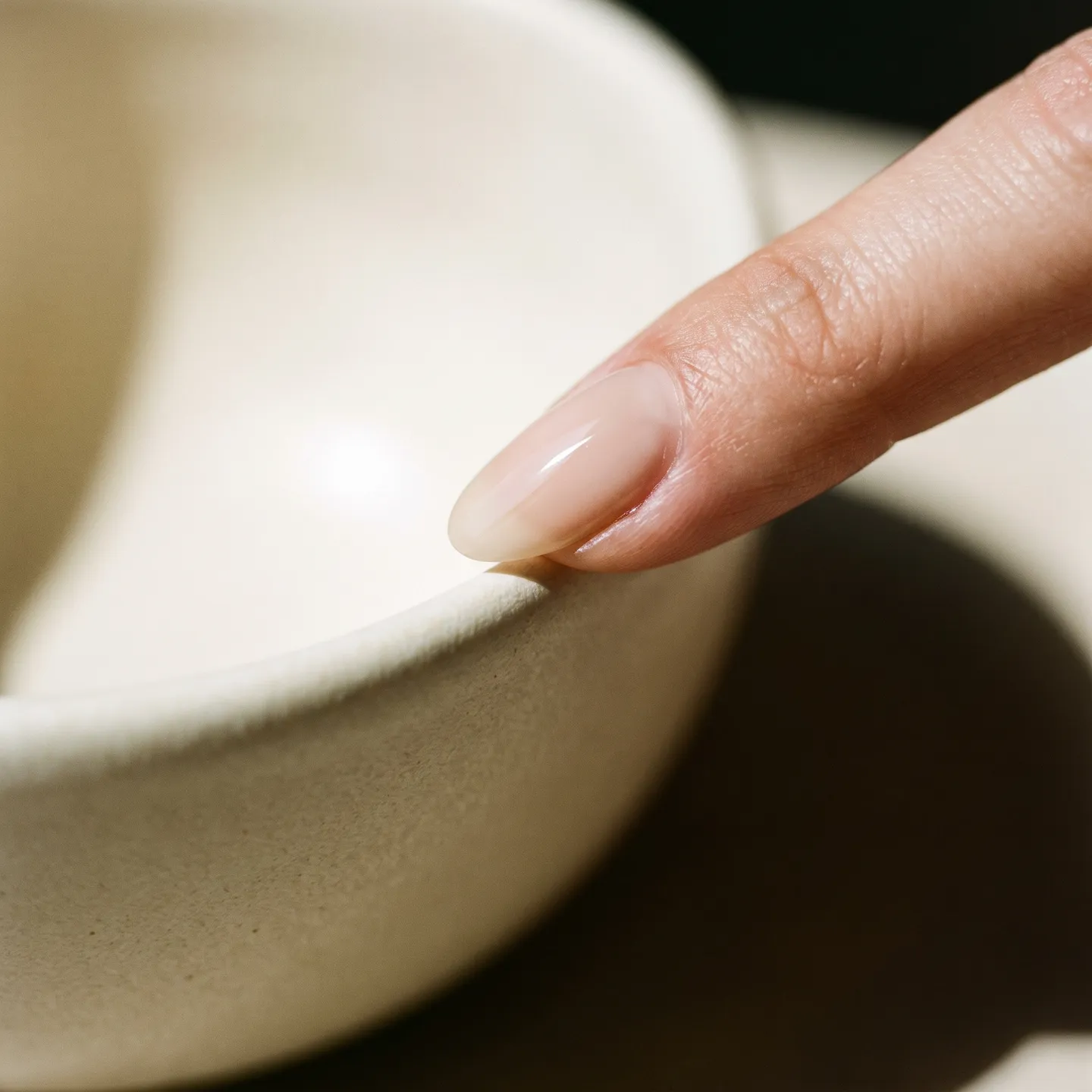 Macro detail of a glossy nude nail touching the rim of an ivory ceramic bowl